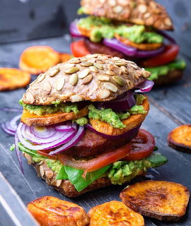 Two tall gourmet veggie burgers with seeded buns, vegetables, and plantain chips on slate