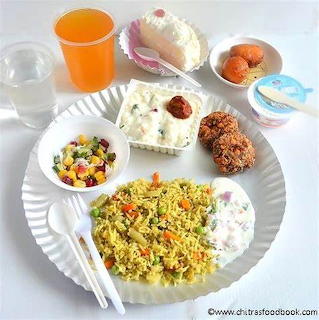 Paper plate with Indian meal: spiced rice, raita, fried snacks, fruit salad, sweets, juice glass, and yogurt cup
