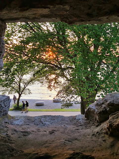 View from a rock cave opening toward a beach with trees and sunset over water