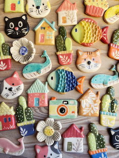Colorful glazed ceramic magnets shaped like cats, fish, houses, cacti, flowers, and cameras on wood