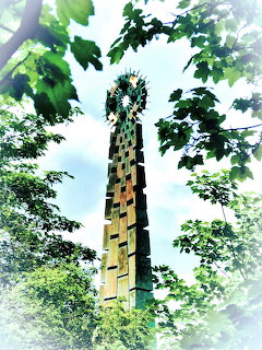 Tall Celtic cross sculpture with mosaic tiles surrounded by green trees viewed from below