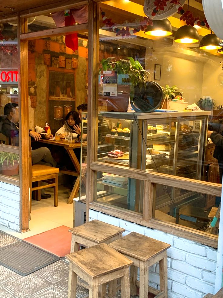 Exterior view of a small cafe with wooden stools, glass pastry display, and customers seated inside
