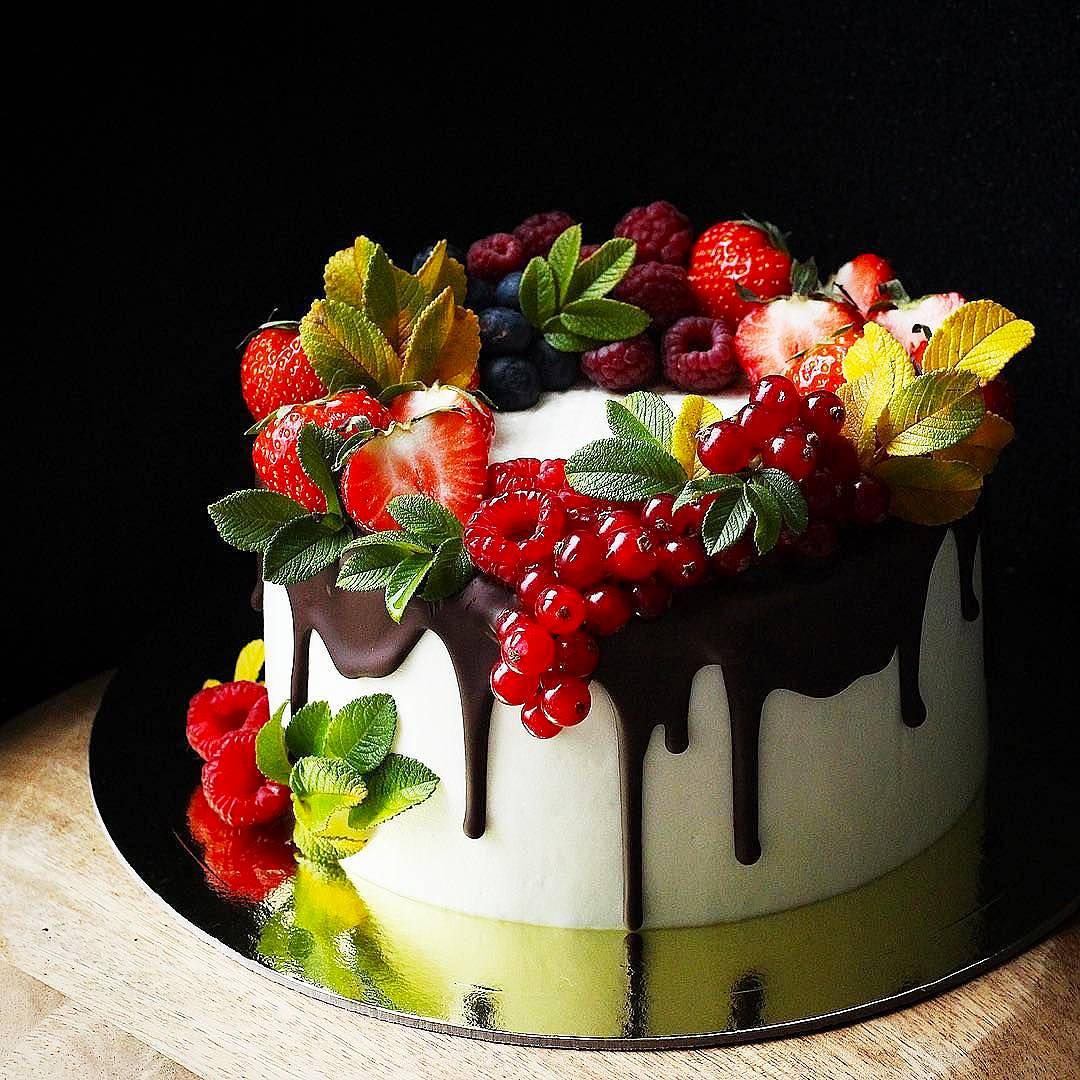 White cake with chocolate drip decorated with strawberries, kiwi, red currants, and yellow orchids on wooden surface