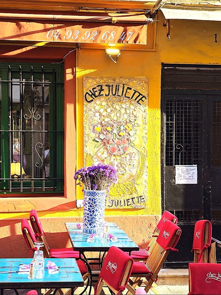 Yellow French cafe exterior with red chairs, blue tables, and floral mosaic sign
