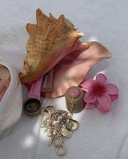 Pink conch shell beside lip gloss, gold jewelry and a pink flower on white fabric