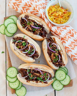 Three spicy chicken sandwiches on baguette rolls with red cabbage, cucumber slices, and corn salad on wooden board