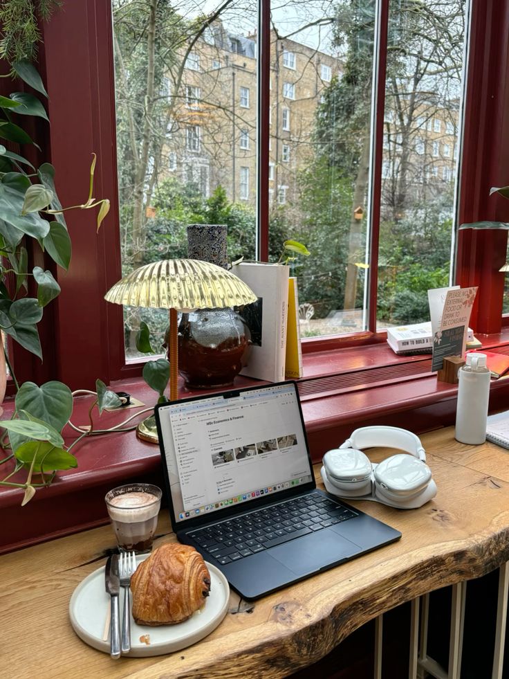 Laptop, croissant, headphones, and a decorative lamp on a wooden table by a large cafe window