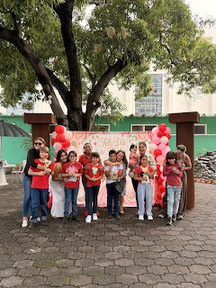 Group of children posing with heart crafts in front of a pink banner and balloons at a school courtyard