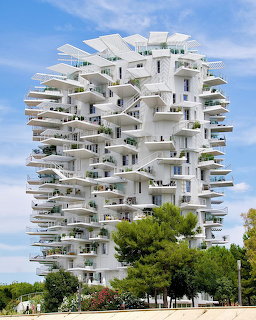 Modern white apartment building with stacked irregular balconies surrounded by green trees under blue sky