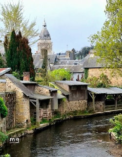 Stone washhouses beside a river in a Normandy village with rooftops and a church tower in the background
