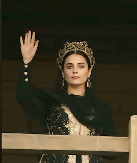 Woman in ornate historical royal dress and crown raising her hand indoors