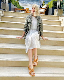 Blonde woman in white dress and camo jacket sitting on wide outdoor stone steps smiling