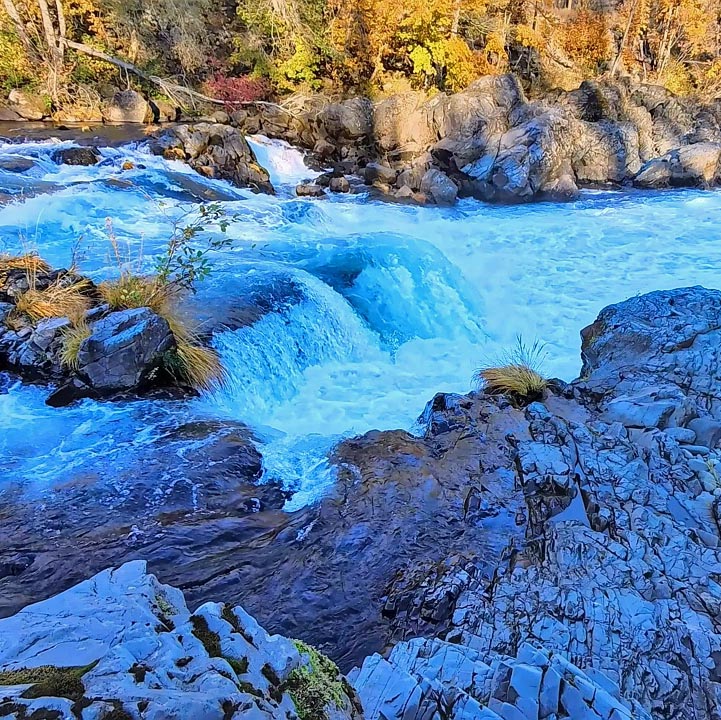 Blue rushing waterfall flowing over dark rocks with autumn-colored trees along the banks