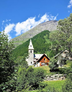 White church and wooden chalet in mountain valley with forested slopes and rocky peak under blue sky