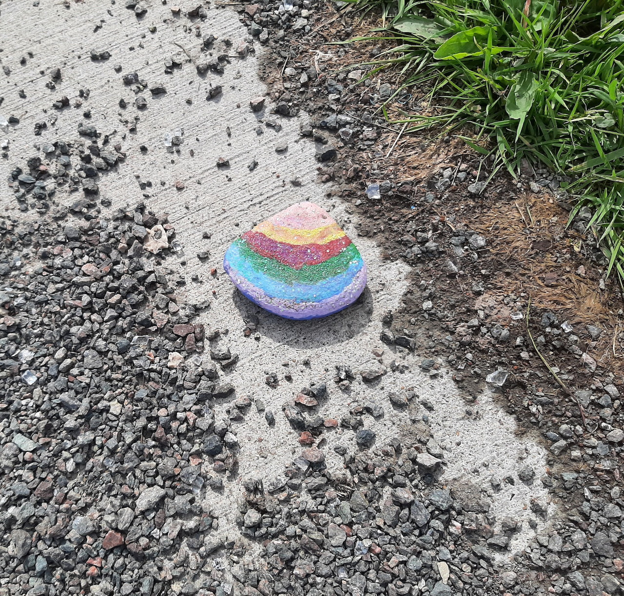 Rainbow-painted rock on concrete beside gravel and grass