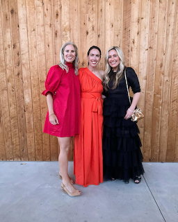 Three women posing in red, orange, and black dresses in front of a wooden wall.