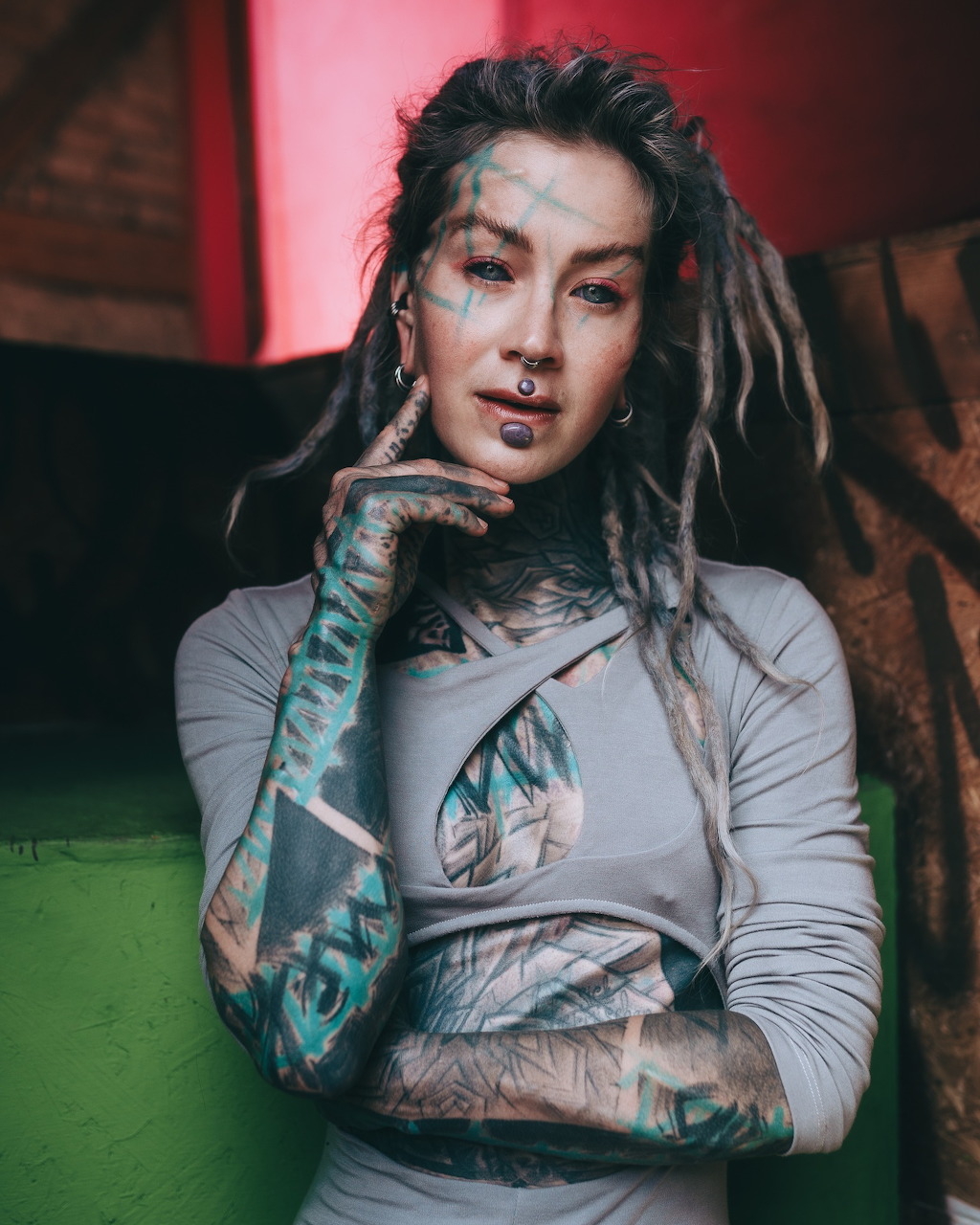 Tattooed woman with dreadlocks and facial piercings in a grey top, arms crossed