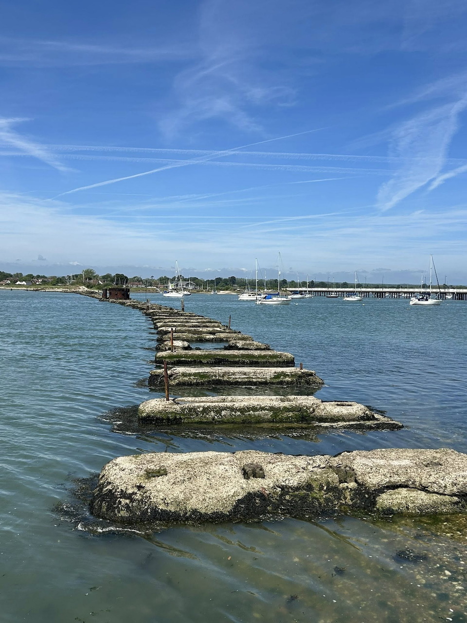 Old stone piers extending across harbor water with boats and shoreline in background under blue sky