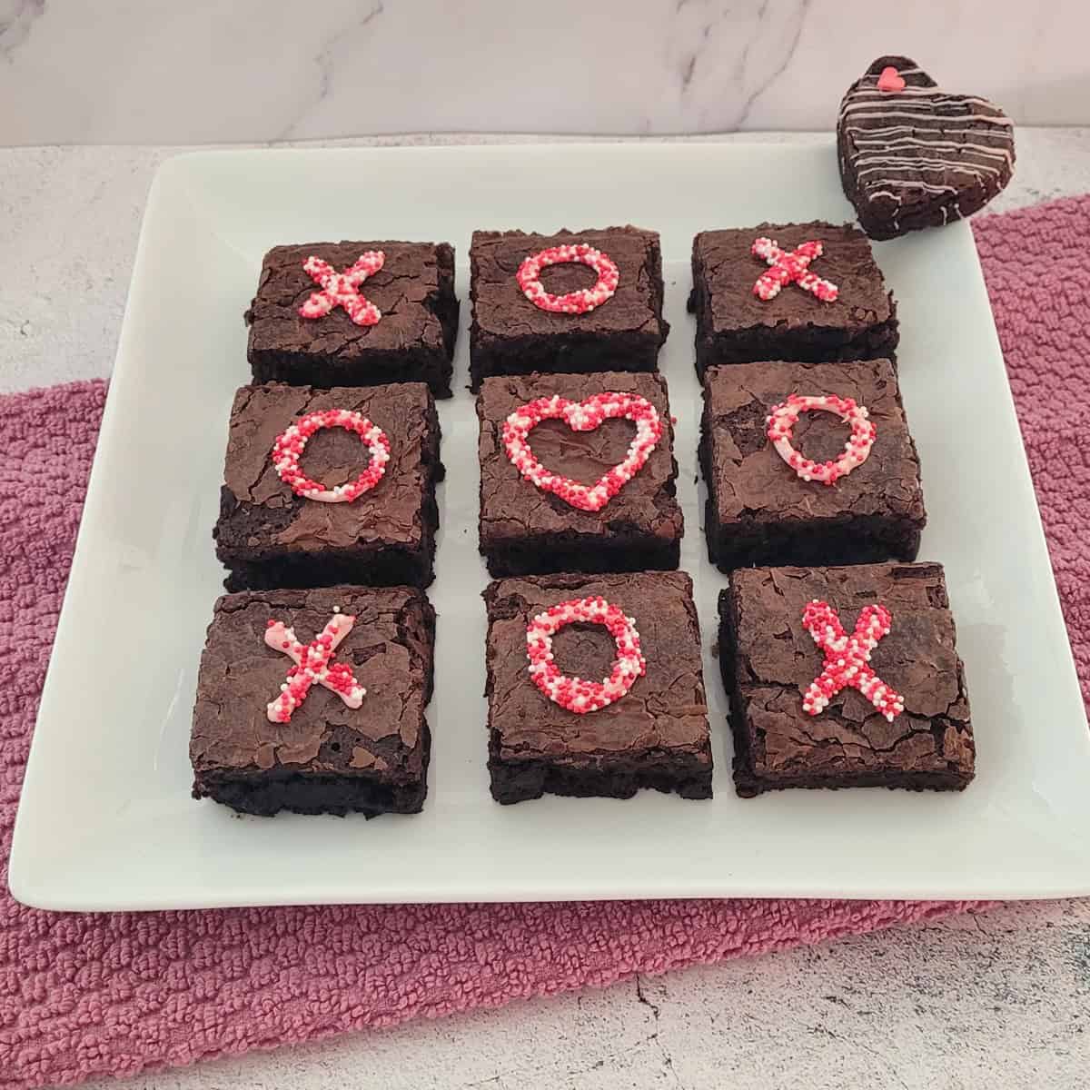 Nine chocolate brownies with pink X's, O's, and heart decorations arranged on white plate