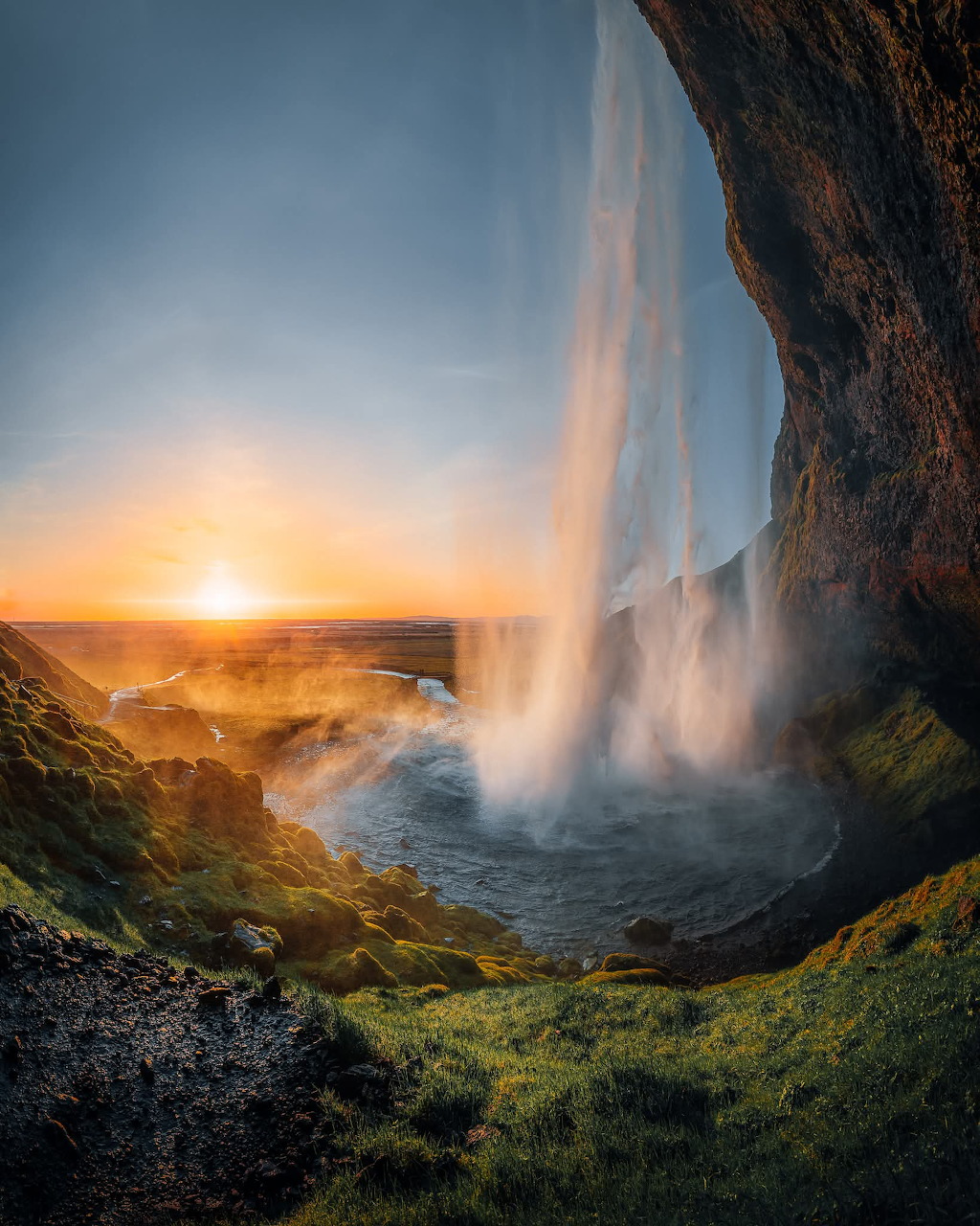 View from behind an Icelandic waterfall at sunset, with golden light and misty cascade over green cliffs