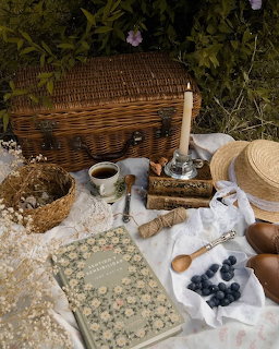 Cottagecore picnic setup with wicker basket, candle, straw hat, book, blueberries, and tea cup on grass