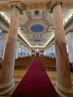 Ornate neoclassical hall with tall columns, decorated ceiling, red carpet, and wooden seating