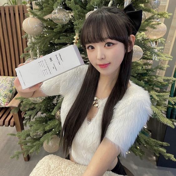 Woman with long dark hair wearing cat ears and white vest, holding white box near Christmas tree