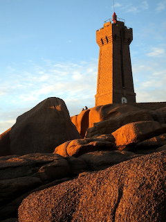 Orange lighthouse tower on rocky coastal outcrop against blue sky at golden hour