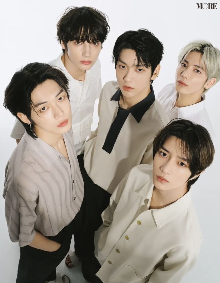 Five young men from K-pop group TXT posing together in cream and black outfits, shot from above