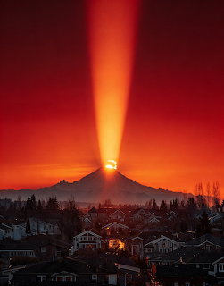 Mount Rainier at sunrise with golden light beam shooting upward through red-orange sky over homes and trees