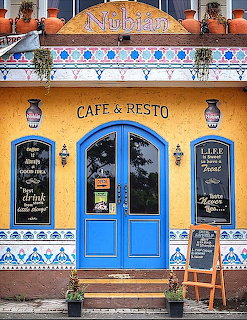 Ornate cafe entrance with blue double doors, yellow walls, decorative tiles, hanging lanterns, and sidewalk chalkboard menus