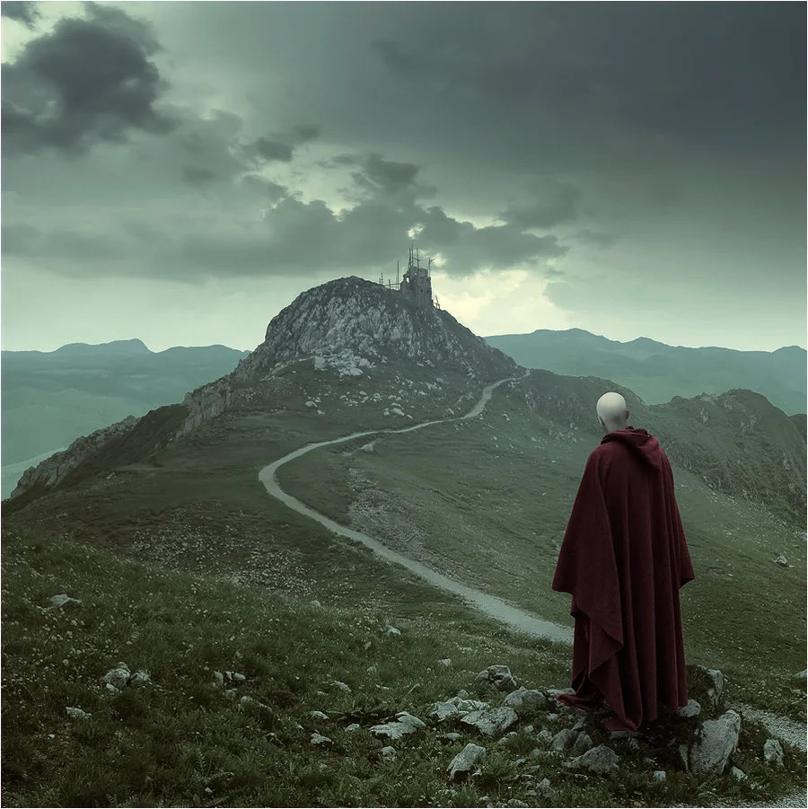 Cloaked figure on a mountain looking toward a winding path and small tower beneath dark clouds