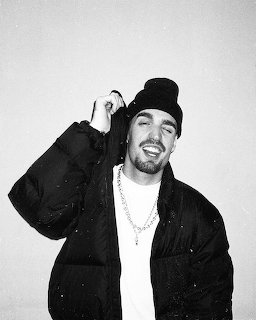 Black and white photo of a smiling man in a black beanie and puffer jacket with a chain necklace