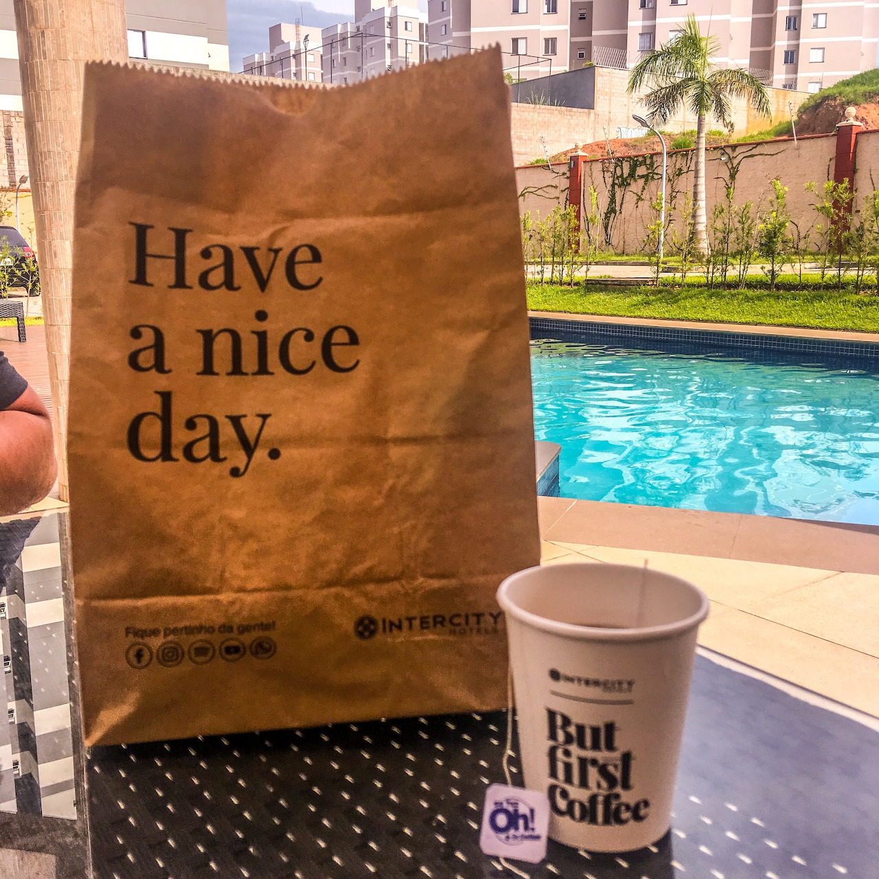 Paper bag reading 'Have a nice day' and coffee cup on poolside table with turquoise pool and palm trees behind