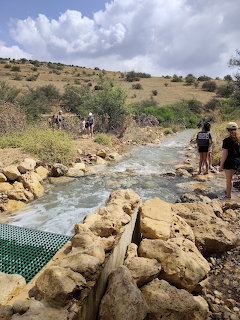Hikers walking beside a flowing stream with rocks and green hills in the background
