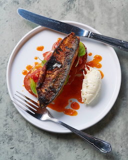Grilled sardine on white plate with orange sauce, grens, and cream alongside knife and fork