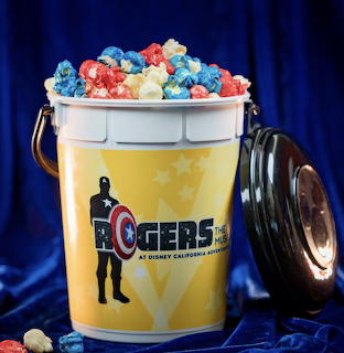 Rogers: The Musical themed popcorn bucket filled with red, white, and blue popcorn on blue satin background