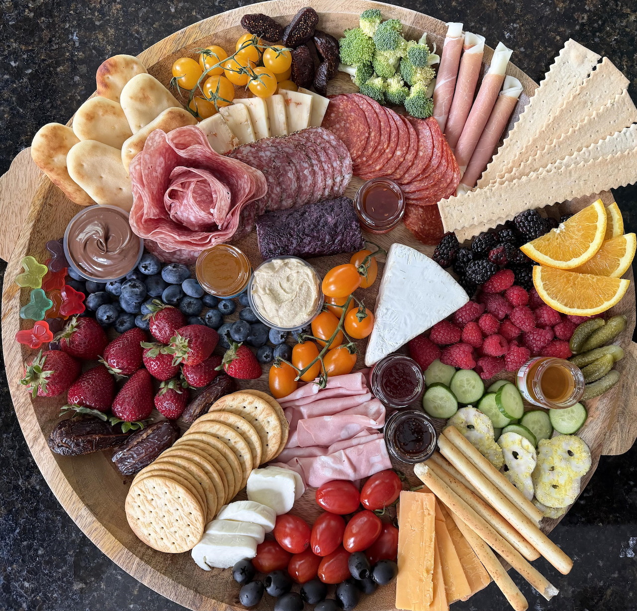 Round charcuterie board with meats, cheeses, crackers, berries, grapes, citrus, and breadsticks