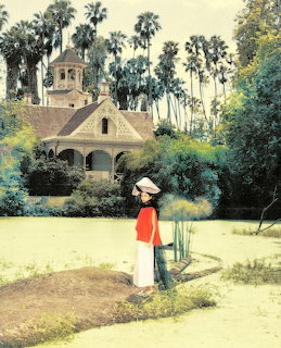 Woman in red jacket and white skirt standing in garden path before historic Spanish-style house with palm trees