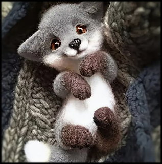 Cute needle-felted gray and white fox toy with brown paws tucked into a knitted sweater pocket
