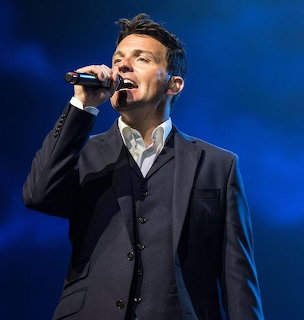 Male singer in a dark suit singing into a microphone on a blue lit stage