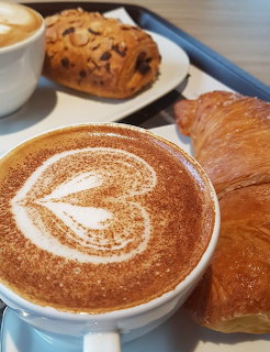 Cappuccino with heart-shaped latte art next to croissants and a chocolate almond pastry on a table
