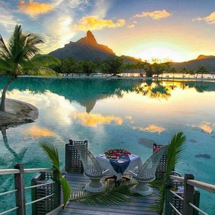 Table for two by a tropical lagoon at sunset with palm trees, mountain, and reflections