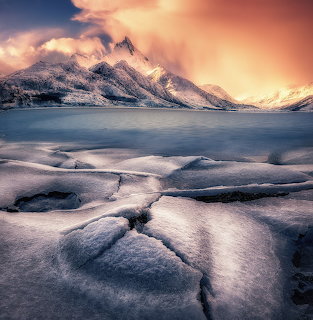 Snowy shoreline with calm water and jagged mountains at sunset in the Lofoten Islands, Norway