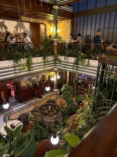 Multi-level indoor cafe atrium with lush green plants, stone fountain, and diners on upper balcony