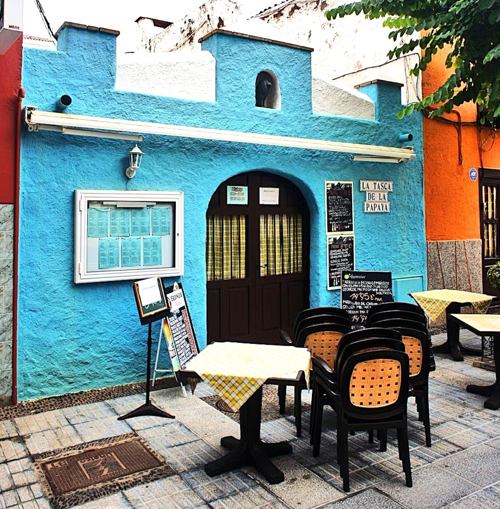 Bright blue cafe storefront with arched entrance, menu board, and outdoor seating on cobblestone patio