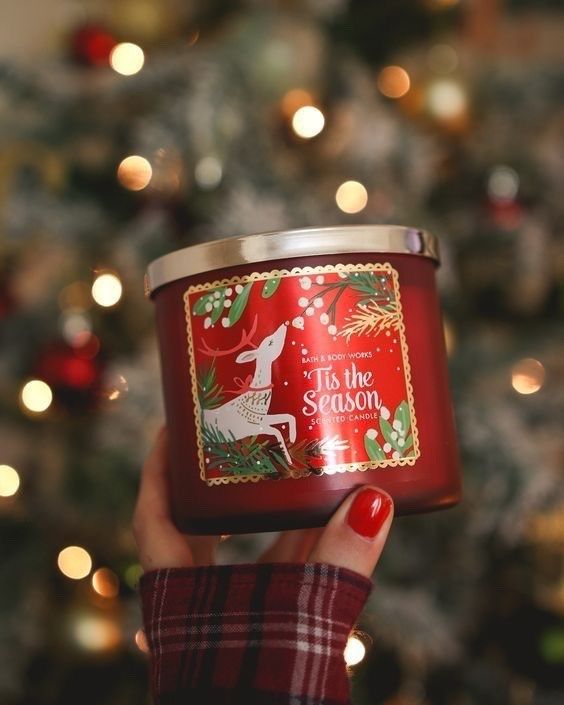 Hand holding a red Christmas candle labeled Tis the Season in front of a lit Christmas tree