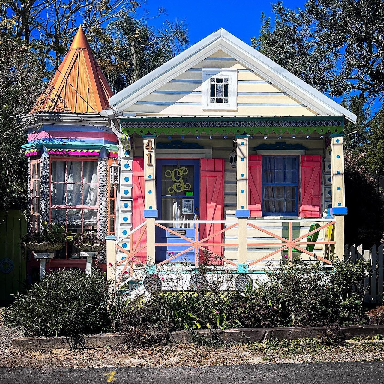 Small white cottage with pink doors, colorful chairs on porch, turret with copper roof, surrounded by shrubs