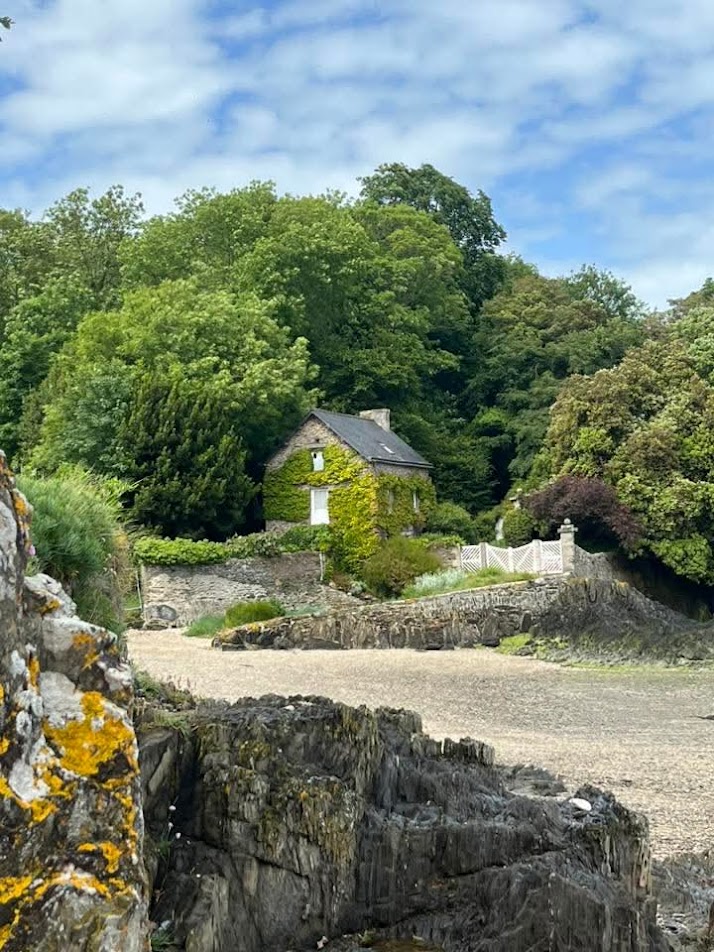 Stone cottage surrounded by dense green foliage with sandy clearing and blue sky