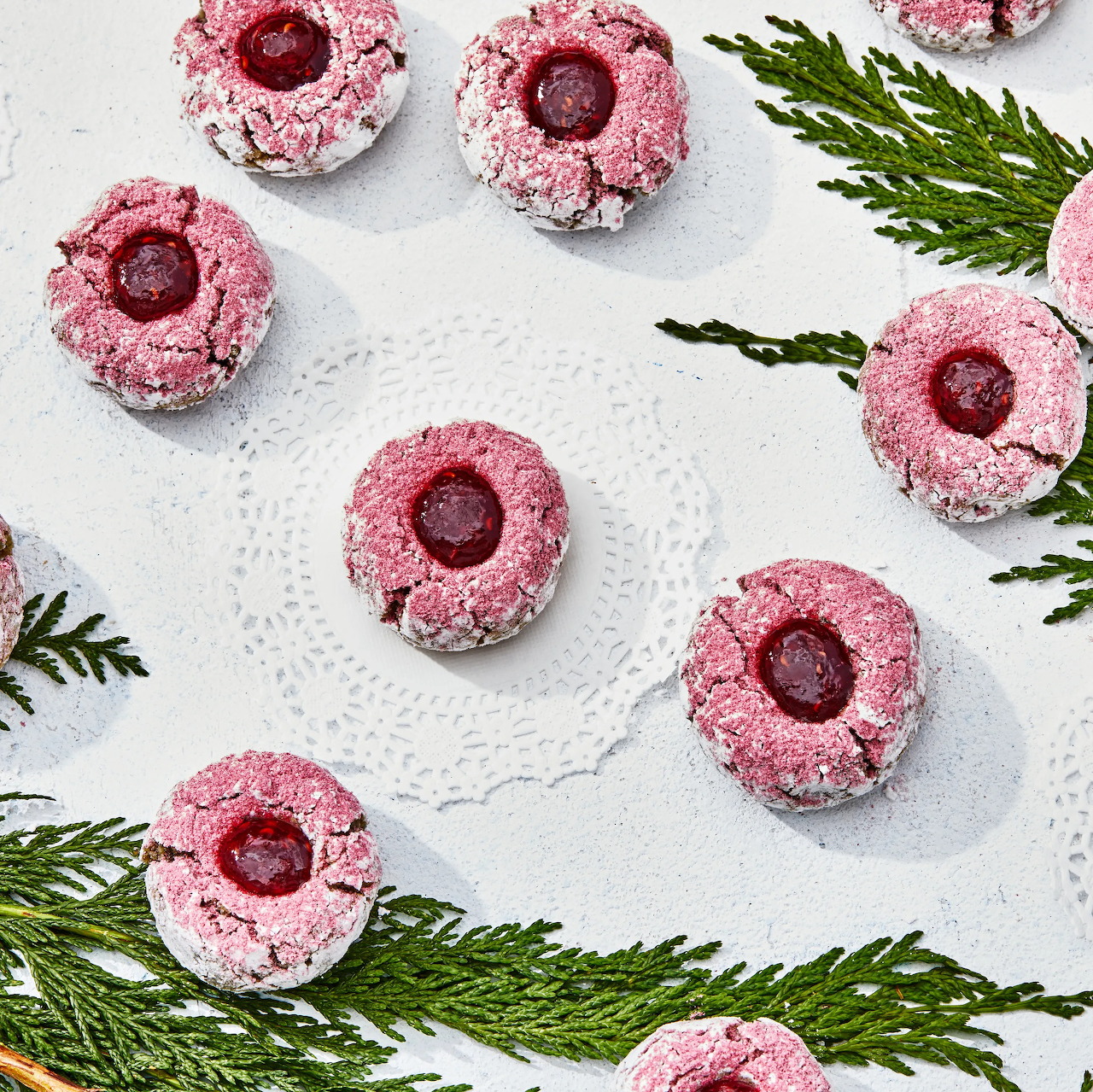 Pink thumbprint cookies with raspberry jam centers on white lace doily with evergreen sprigs